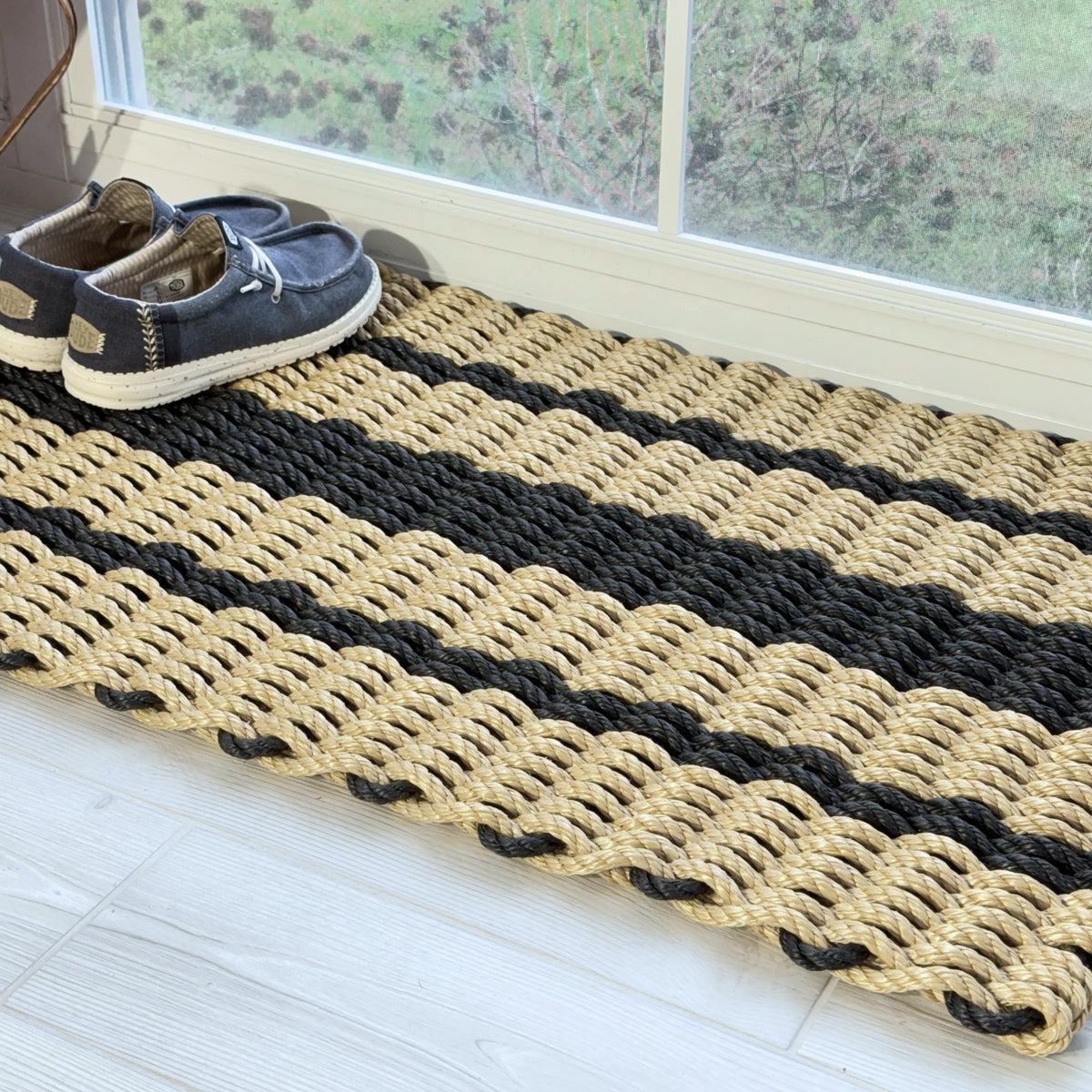Angled view of a tan and black lobster rope doormat beside a screened window with blue boat shoes on one corner.