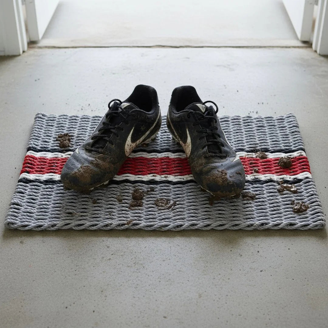 Limited-edition Sun and Salt Edit seasonal rope doormat featuring a Ohio State football themed doormat, hand-woven in Southport, North Carolina, styled with muddy cleats sitting on top of the gray, red, white and black striped all season, outdoor, durable lobster rope doormat.