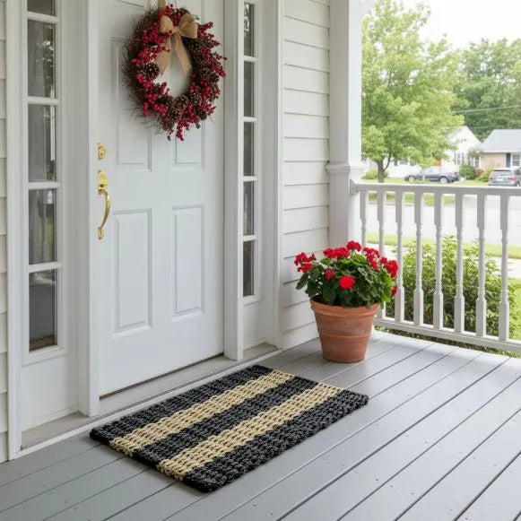 Rope Doormat with black and Tan Classic stripe styling sitting on front porch with a white door, wreath, and potted plant.