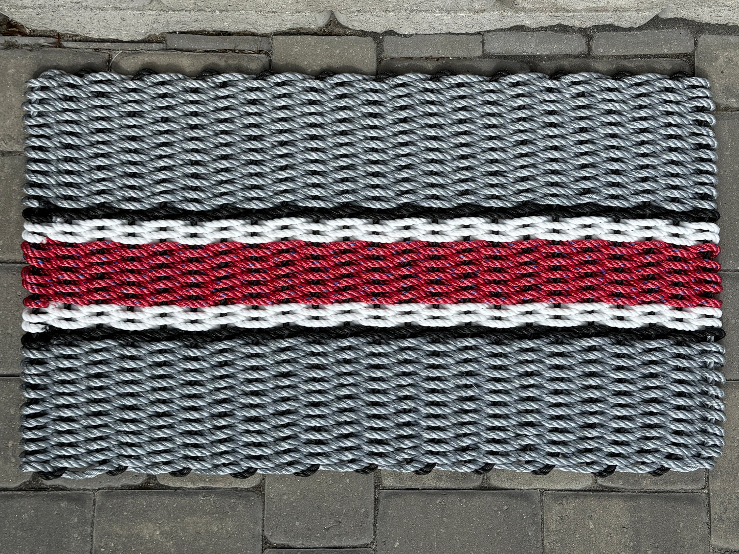Doormat with a woven pattern featuring gray, red, and black colors on a stone floor.