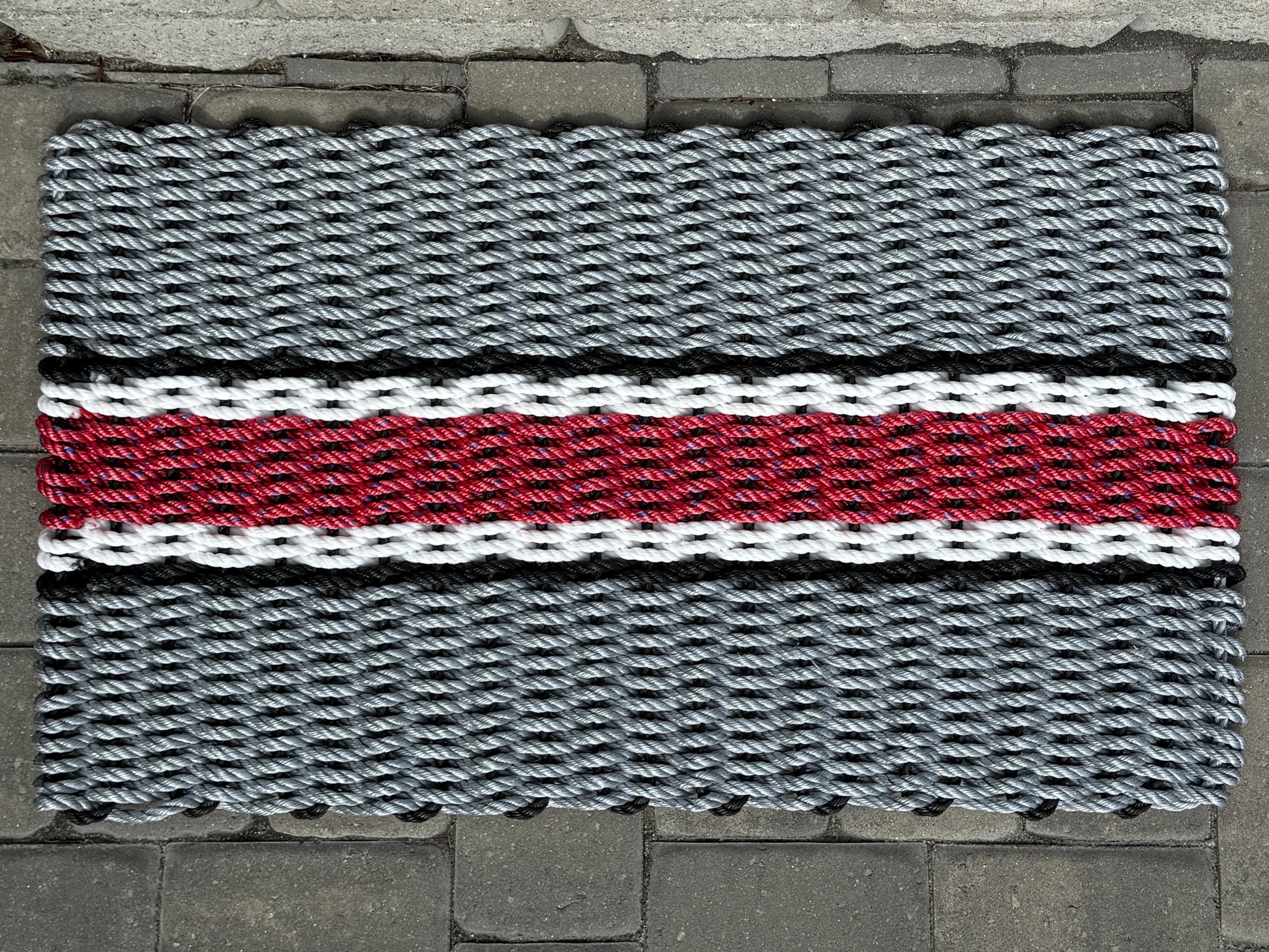 Doormat with a woven pattern featuring gray, red, and black colors on a stone floor.
