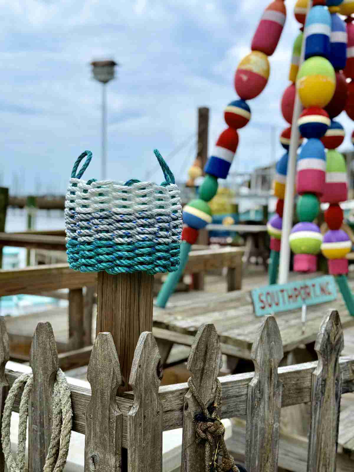 All-weather, durable coastal rope basket teal gray and white on post in downtown Southport NC with buoys and other coastal decor in the background