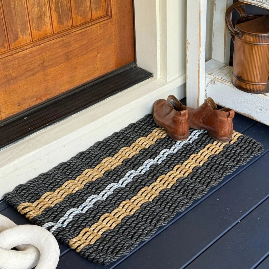 Handwoven striped doormat with black, tan and gray stripes sitting on a front porch with leather shoes on it.