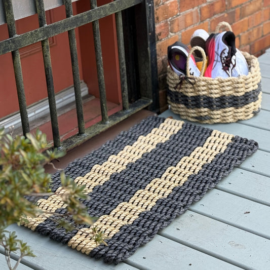 Textured black and tan outdoor welcome doormat with striped pattern on a coastal cottage farmhouse porch with a matching lobster rope basket.  